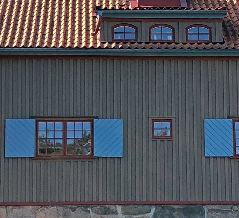Detalj av trähus med grågrön fasad, röda fönster och detaljer samt blå fönsterluckor
