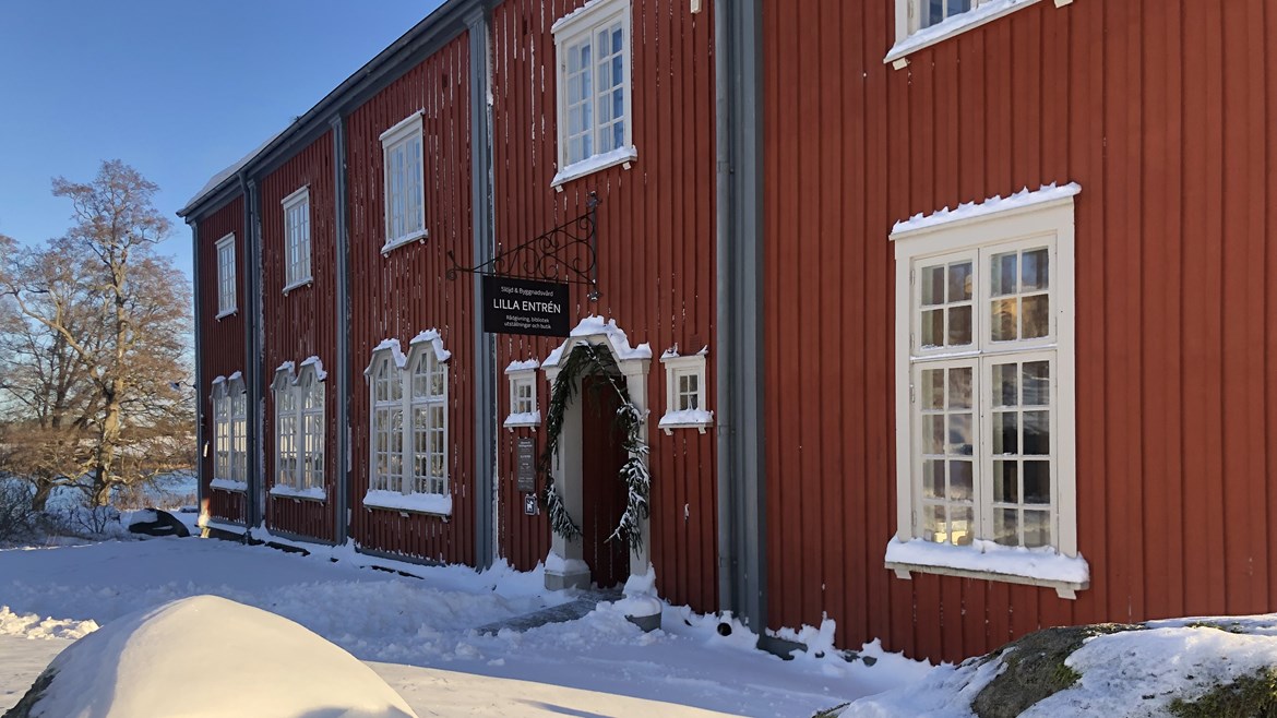 Ett stort rödbrunt hus med grå och vita detaljer och en butiksentré med granris. En kall vinterdag med snö på marken och på fönsteröverstycken