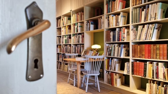 Ett dörrblad med ett äldre dörrhandtag i förgrunden och en vägg med fyllda bokhyllor i bakgrunden.